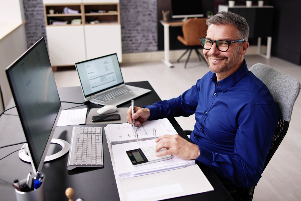 Ein Mann sitzt vor mit einem Taschenrechner vor einem PC und beschäftigt sich mit Buchhaltung.